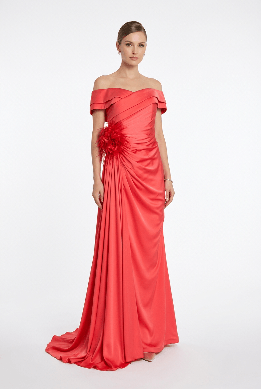 A woman modeling a floor-length off-the-shoulder gown in bright coral-red satin, with ruched draping at the waist and a large matching fabric flower detail, photographed against a clean light gray studio background.