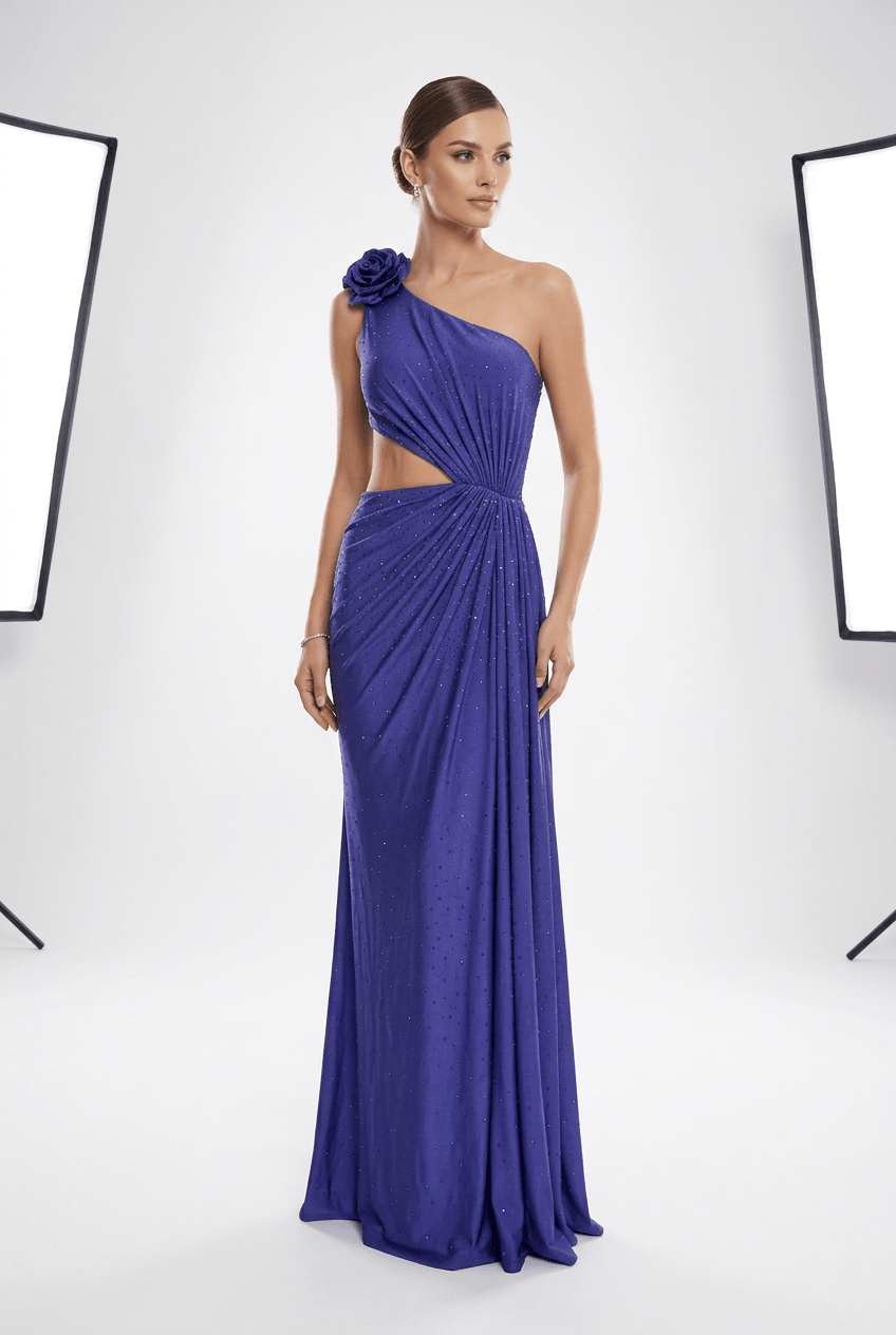A woman models a floor-length one-shoulder gown in a studio setting. The dress is a rich royal blue with a draped, gathered design, a side cutout, and a large fabric flower detail on the shoulder.