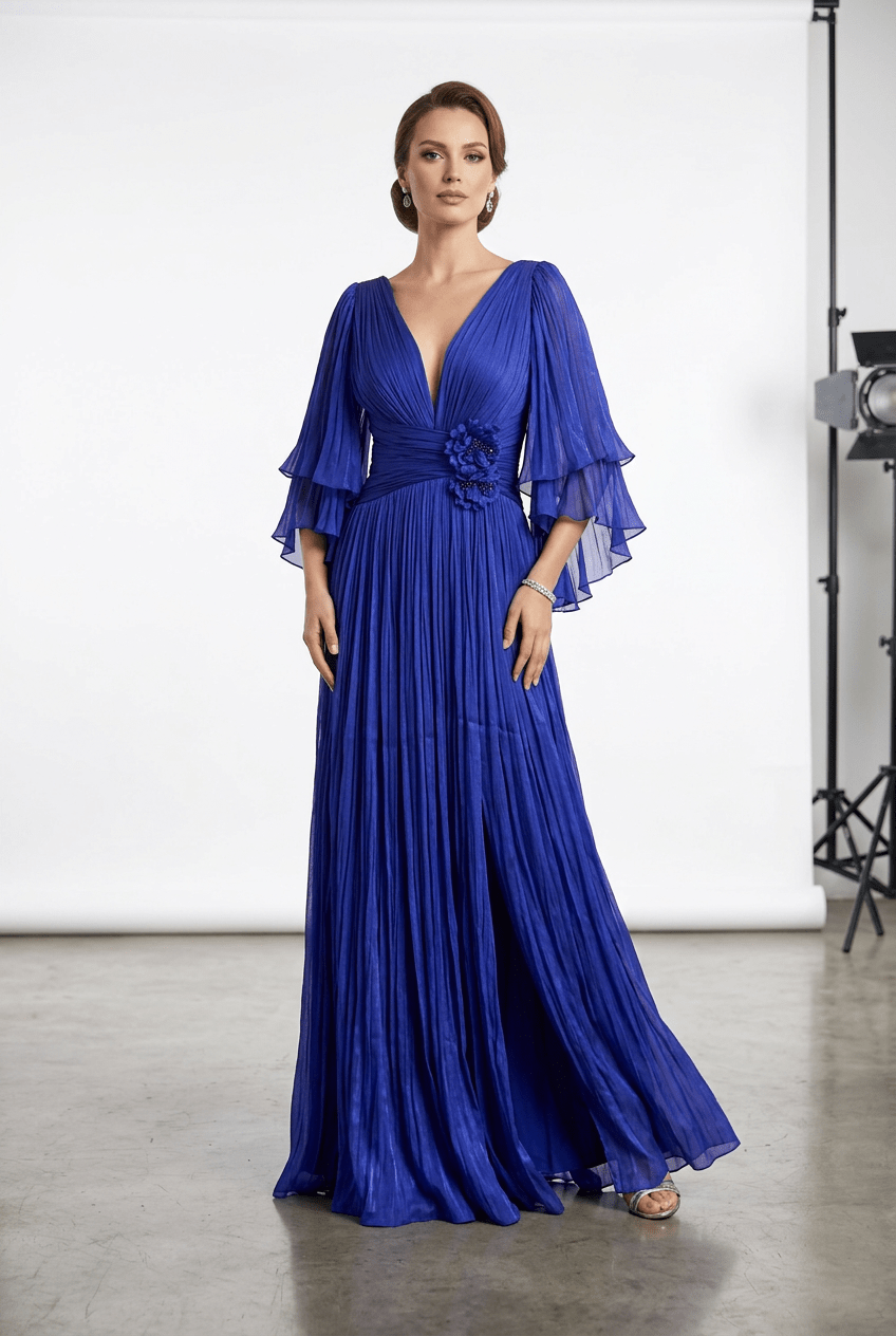 A woman modeling a floor-length royal blue pleated gown with a deep V-neck, layered flutter sleeves, and a floral waist detail, photographed in a studio setting.