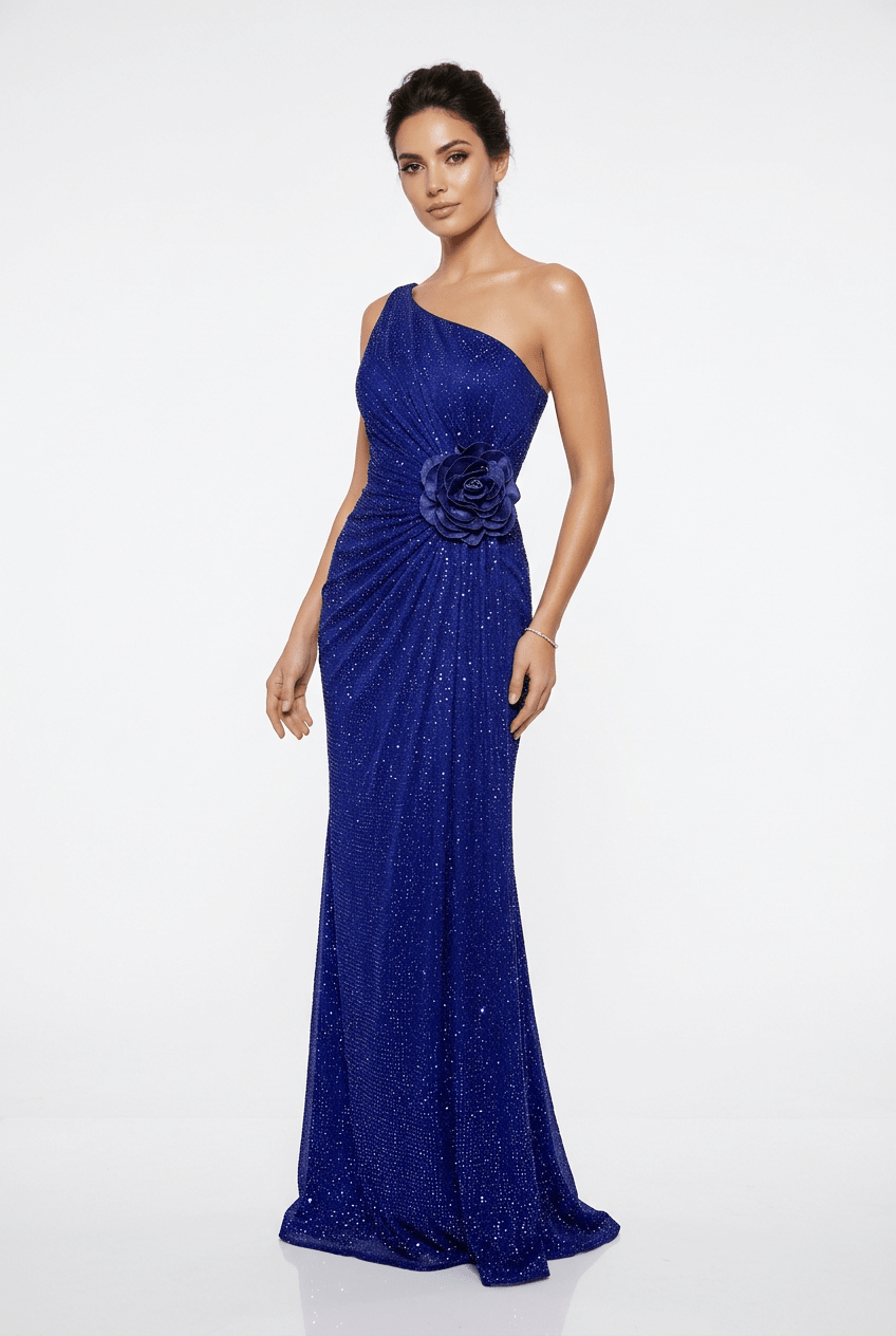 A woman modeling a floor-length royal blue one-shoulder evening gown with sequins, a draped waist, and a large fabric flower detail at the waist, photographed against a plain white studio background.
