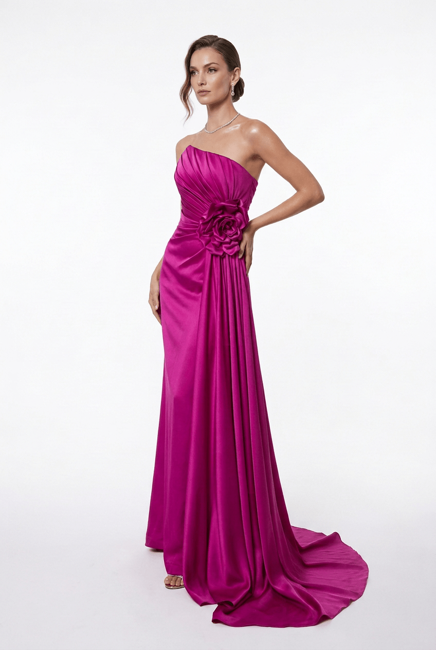 A woman modeling a bright fuchsia magenta strapless formal gown with a draped waist detail and a long train, photographed against a plain white studio background.