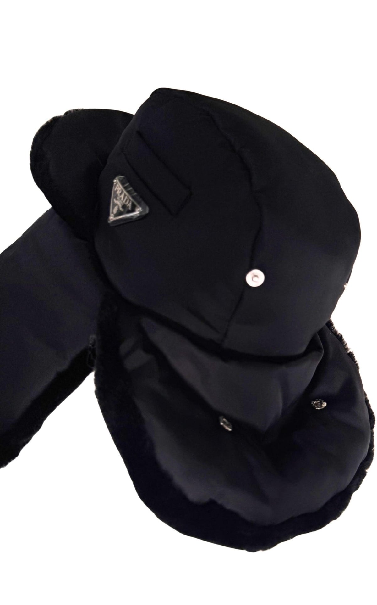 A black winter trapper hat with attached ear flaps and a chin guard, featuring a small silver-tone triangular logo patch and a snap button on the crown, set against a plain white background.