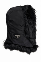 A black Prada fur-trimmed hat featuring a black nylon body, a gold-tone triangular Prada logo on the front, and a fluffy black fur trim at the top and bottom, shown against a plain white background.