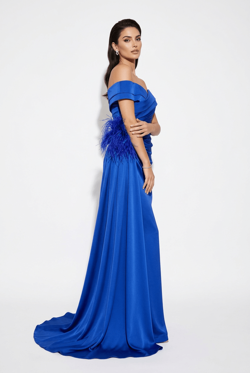 A woman modeling an off-the-shoulder royal blue evening gown with a feathered waist detail, photographed against a plain white studio background.