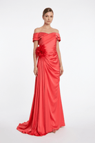 A woman modeling a floor-length off-the-shoulder gown in bright coral-red satin, with ruched draping at the waist and a large matching fabric flower detail, photographed against a clean light gray studio background.