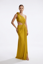 A woman modeling a floor-length yellow one-shoulder evening gown with a draped cutout bodice, floral shoulder detail, and gathered skirt against a plain light gray studio background.