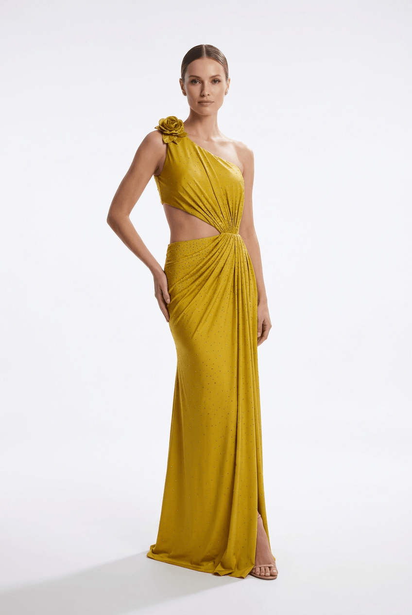 A woman modeling a floor-length yellow one-shoulder evening gown with a draped cutout bodice, floral shoulder detail, and gathered skirt against a plain light gray studio background.