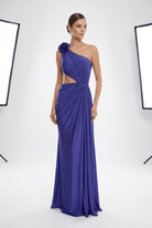 A woman models a floor-length one-shoulder gown in a studio setting. The dress is a rich royal blue with a draped, gathered design, a side cutout, and a large fabric flower detail on the shoulder.