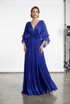 A woman modeling a floor-length royal blue pleated gown with a deep V-neck, layered flutter sleeves, and a floral waist detail, photographed in a studio setting.
