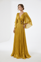 A woman models a floor-length pleated gown in a rich mustard yellow color. The dress features a deep V-neckline, a decorative embroidered bodice, and dramatic, flowing bell sleeves.