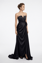 A woman modeling a strapless black satin evening gown with a sweetheart neckline, embellished bodice, draped waist, and floor-length train against a light gray studio background.