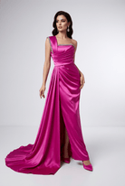 A woman modeling a floor-length magenta satin gown with a draped one-shoulder neckline, gathered waist, and a slight train on a light gray studio background.