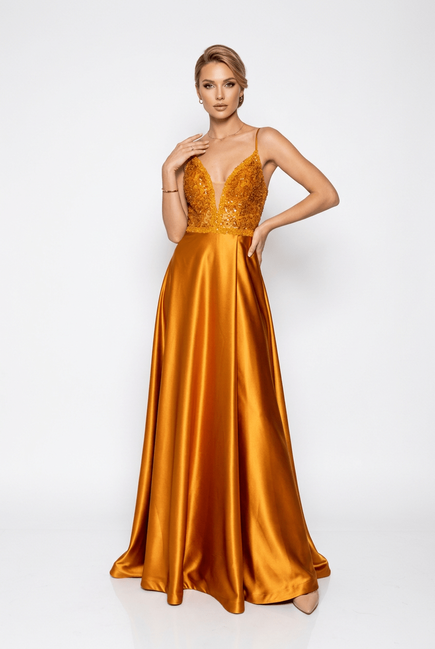 A woman modeling a floor-length satin gown in a rich golden-amber color. The dress features a beaded, embellished V-neck bodice with thin straps and a flowing skirt, photographed against a clean white studio background.