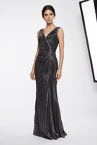 A woman modeling a floor-length, sleeveless evening gown with a deep V-neckline and a wrap-style front overlay, in a dark metallic gray or black fabric with a subtle shimmer. The dress features a gathered, pleated skirt.