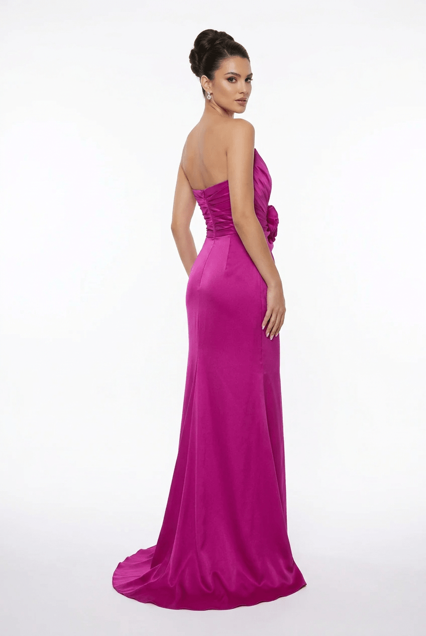 A woman modeling a strapless magenta formal gown with a gathered waist and a long train against a plain white studio background.