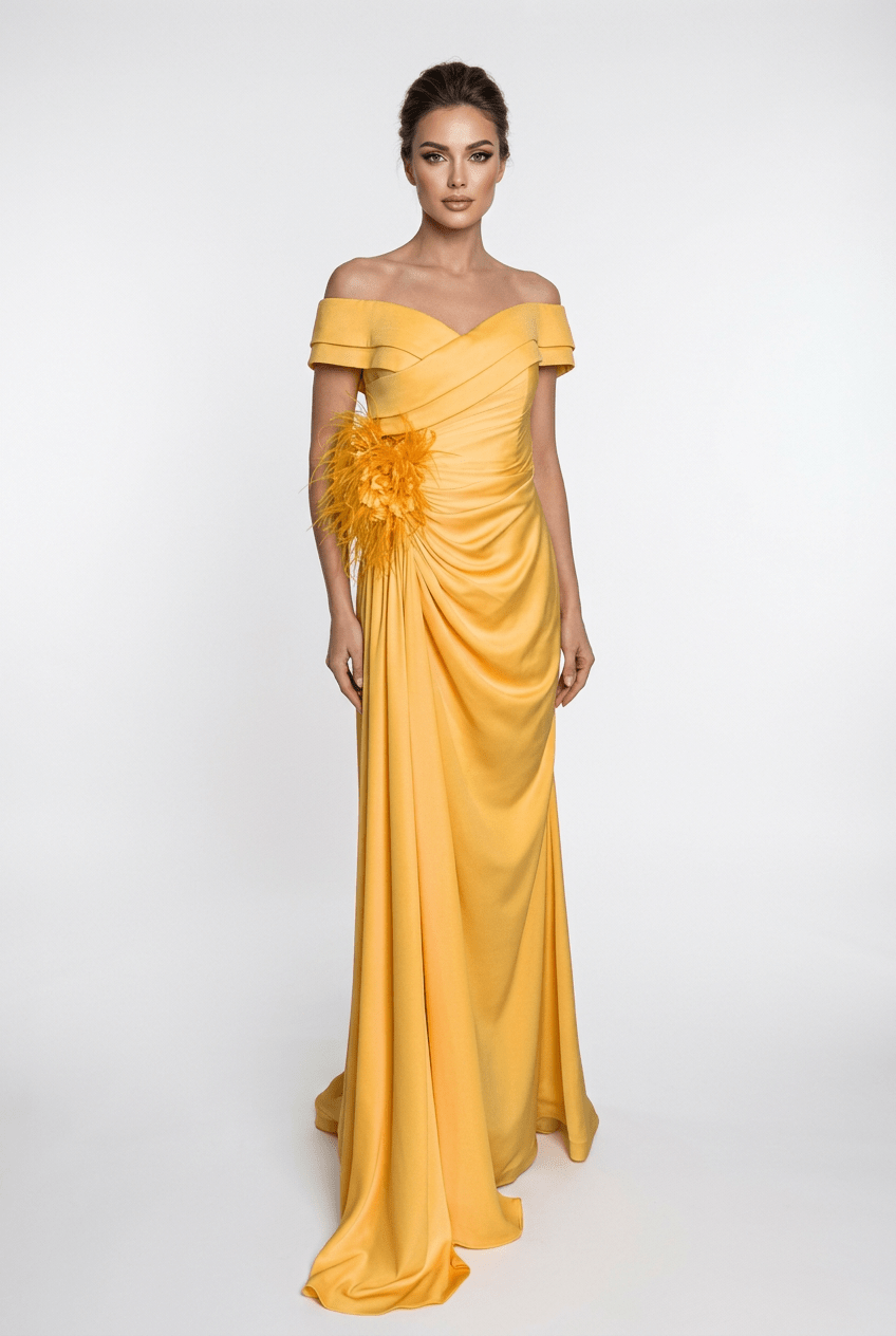 A woman modeling a floor-length off-the-shoulder yellow gown with draped fabric, a gathered waist, and a large feathered floral detail on one side, photographed against a light gray studio background.