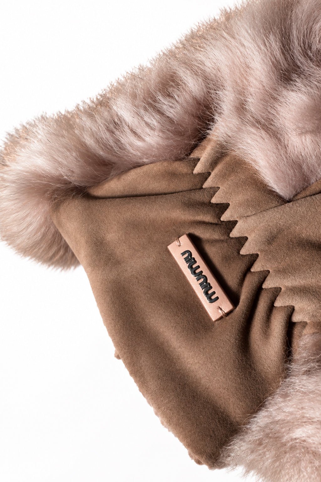 A close-up of a brown suede with a fuzzy pale pink rectangular brand tag reading Miu Miu