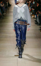 A runway model walks down a fashion show aisle wearing a gray argyle sweater, dark blue Miu Miu leather pants with distressed details, a brown belt, and black boots.