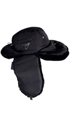 A black winter trapper hat with plush ear flaps and a faux-fur trim, featuring a small silver-tone triangular Prada logo on the front.