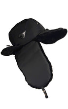 A black Prada winter trapper hat with plush ear flaps and a chin strap, featuring a small triangular logo patch on the side, set against a plain white background.