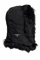 A black Prada fur-trimmed hat featuring a black nylon body, a gold-tone triangular Prada logo on the front, and a fluffy black fur trim at the top and bottom, shown against a plain white background.