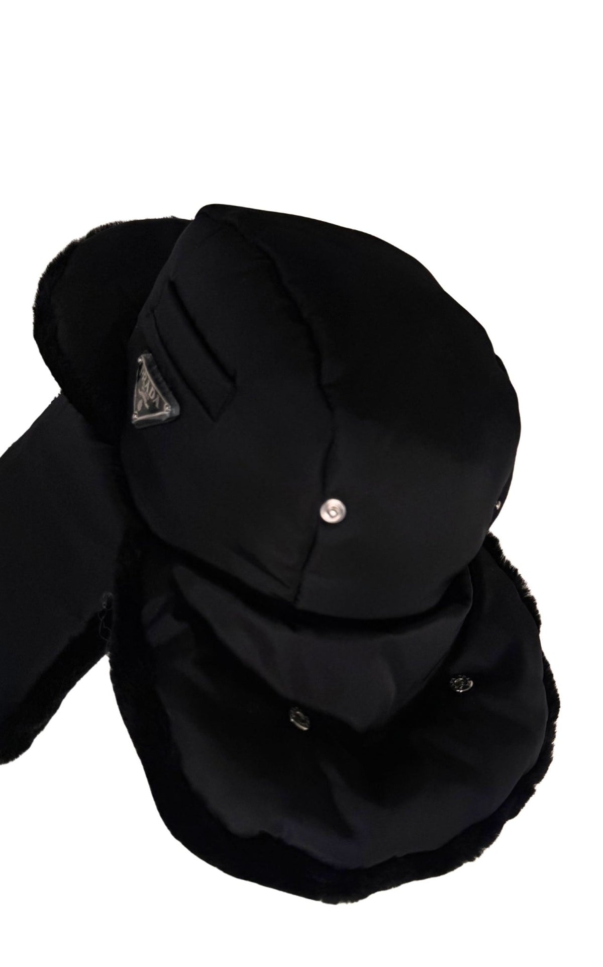 A black winter trapper hat with ear flaps and a chin guard, featuring a small silver metal snap button and a triangular metal logo patch on the side, set against a plain white background.