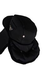 A black winter trapper hat with ear flaps and a chin guard, featuring a small silver metal snap button and a triangular metal logo patch on the side, set against a plain white background.