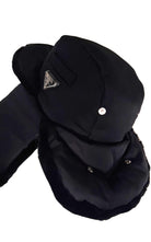 A black winter trapper hat with attached ear flaps and a chin guard, featuring a small silver-tone triangular logo patch and a snap button on the crown, set against a plain white background.