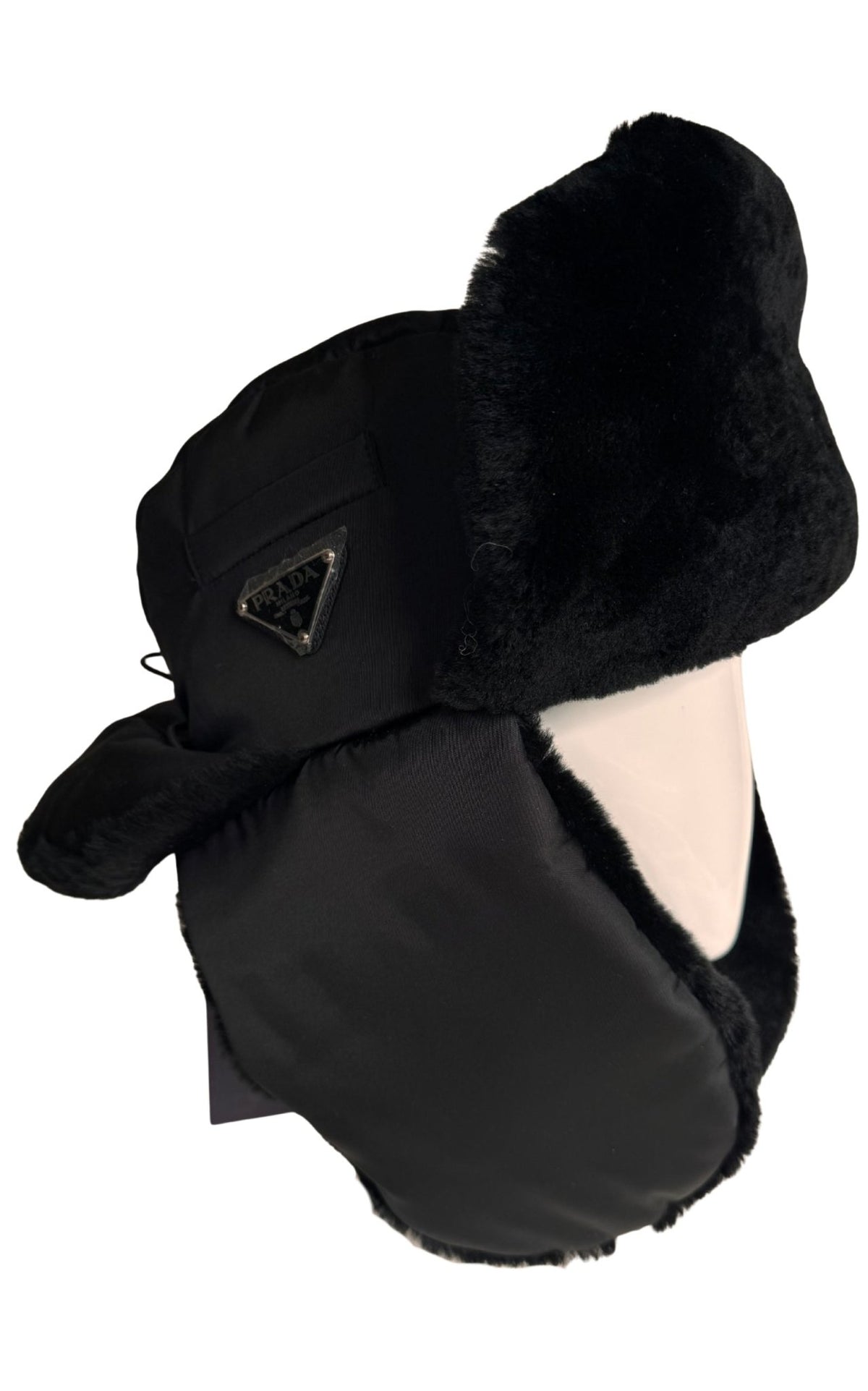 A black Prada winter trapper hat with plush black faux fur ear flaps and a matching neck warmer, featuring a small triangular metal logo badge on the side.