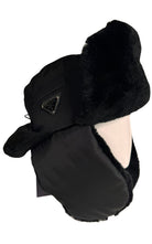 A black Prada winter trapper hat with plush black faux fur ear flaps and a matching neck warmer, featuring a small triangular metal logo badge on the side.