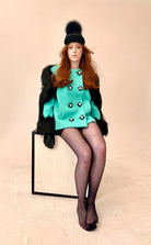 Woman in a turquoise outfit with black fur vest and hat sitting on a box against a beige background